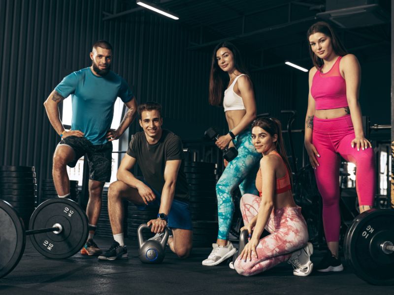 prikaz 5 osoba u teretani na grupnom treningu koji poziraju pored sprava